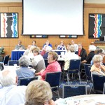 5 people are seated at a panel table at the front of a room, and they and some seated listeners turn to look at a person asking a question